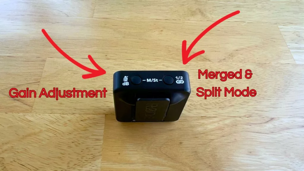 the rode wireless go transmitter flipped on it's side to show the gain button and the merged/split mode button. The transmitter is sitting on a wooden table.